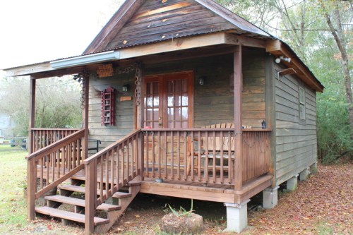 A rustic wooden cabin with a porch, steps, and a welcoming entrance surrounded by greenery.