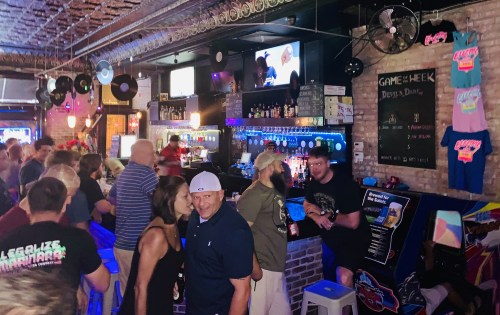 A lively bar scene with a crowd enjoying drinks, colorful decor, and a TV displaying a game in the background.