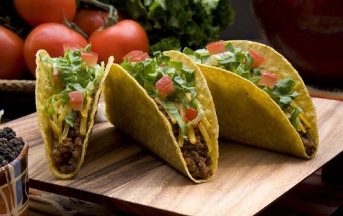 Three tacos filled with lettuce, cheese, and diced tomatoes on a wooden platter, with fresh tomatoes and lettuce in the background.