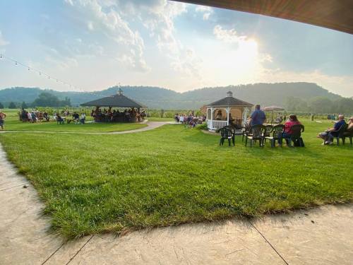 A scenic outdoor gathering with people seated on chairs, gazing at a gazebo and lush green landscape under a cloudy sky.