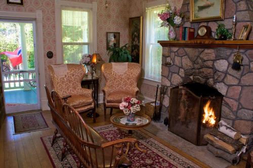 Cozy living room with stone fireplace, vintage chairs, floral decor, and a view of a porch through the window.
