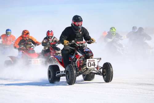 A group of riders on ATVs racing through a snowy landscape, kicking up snow and dust in the air.