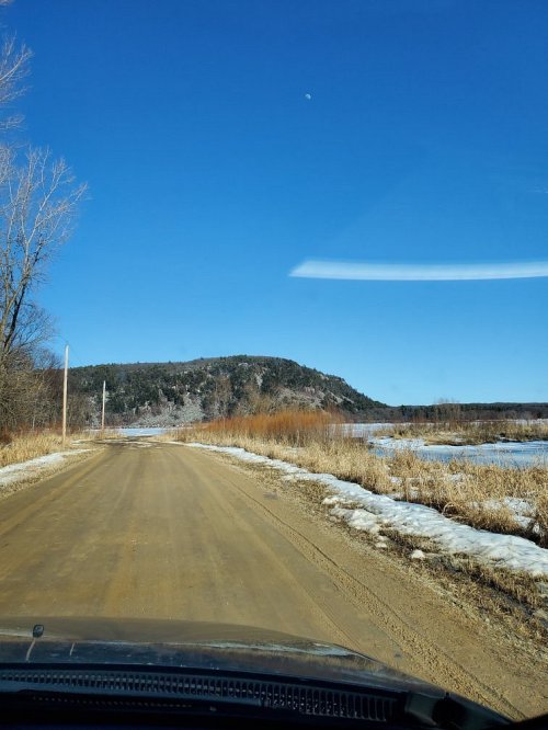 A dirt road leads toward a hill under a clear blue sky, with patches of snow and water visible on the sides.