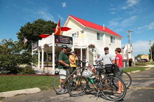 A group of cyclists gathers outside a charming white building with a red roof, enjoying a sunny day.