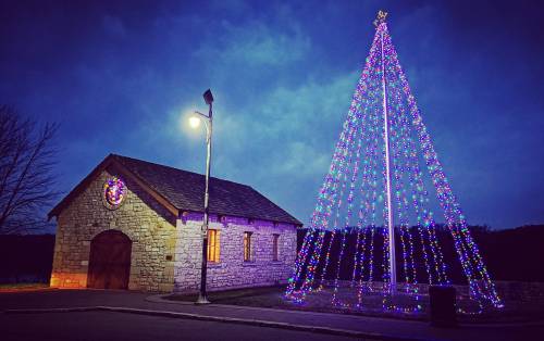 A brightly lit Christmas tree made of lights stands next to a stone building under a cloudy evening sky.