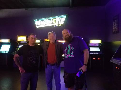 Three men pose together in an arcade, with vintage game machines and a neon sign in the background.