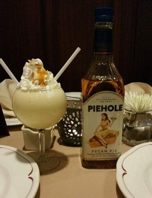 A dessert drink topped with whipped cream and a bottle of Piehole Pecan Pie liqueur on a table setting.