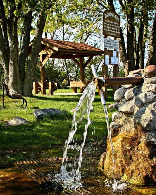 A flowing well with water cascading over rocks, surrounded by greenery and a wooden gazebo in the background.