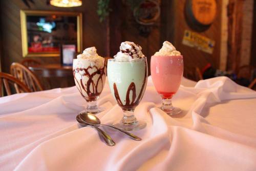 Three colorful milkshakes in tall glasses, topped with whipped cream and chocolate drizzle, on a white tablecloth.