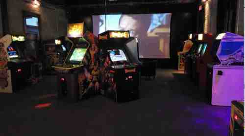 A retro arcade room filled with various arcade machines and a large screen displaying a movie in the background.
