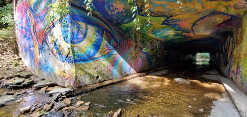 Colorful graffiti featuring a large eye adorns the walls of a tunnel, with a stream flowing through the rocky floor.
