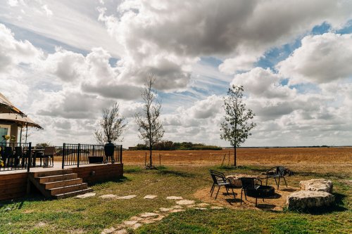 A scenic outdoor area with a wooden deck, chairs, and a fire pit, surrounded by grassy fields and cloudy skies.