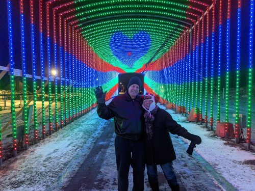 A couple poses joyfully under a colorful light tunnel with red, green, and blue lights in a snowy setting.