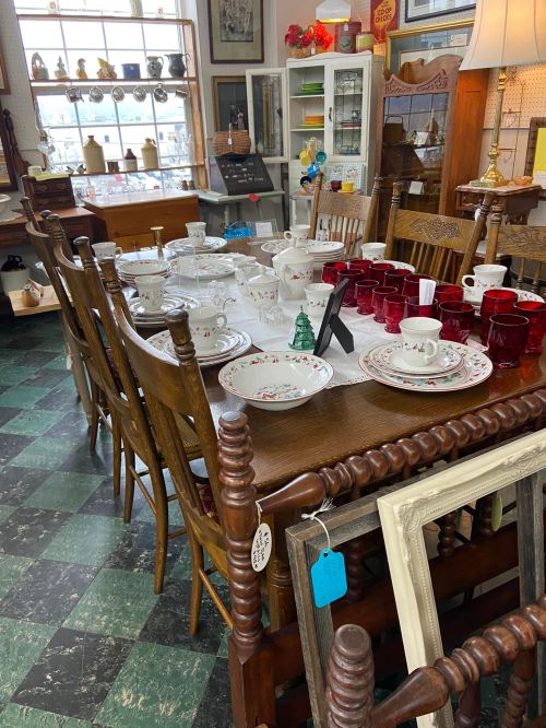 A vintage dining table set with various dishes, cups, and decorative items, surrounded by wooden chairs in a cozy shop.