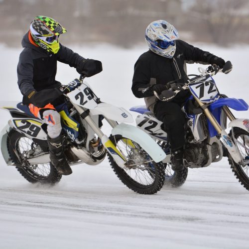 Two motocross riders in winter gear race on snow-covered terrain, showcasing their bikes and skills.