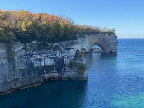 A scenic view of a rocky cliff with an arch, surrounded by calm blue waters and autumn-colored trees.