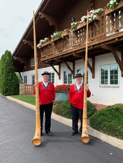 Two men in traditional attire hold large wooden alphorns outside a building with flower boxes and a balcony.
