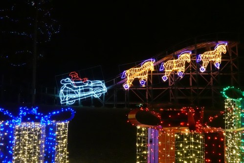 Colorful holiday lights display featuring a roller coaster and illuminated horses, with large gift boxes in the foreground.