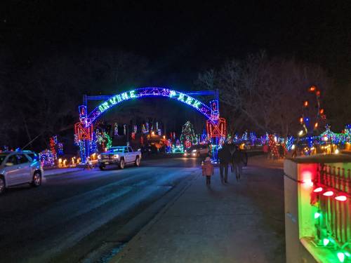 A festive street scene at night, adorned with colorful holiday lights and decorations, with people walking along the road.