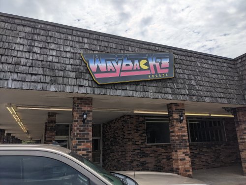 Sign for "Wayback Arcade" on a building with a shingled roof and brick columns under a cloudy sky.