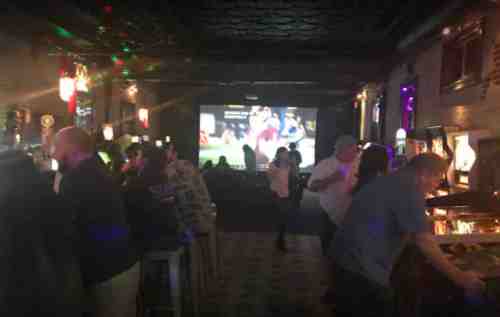 A lively bar scene with patrons socializing, colorful lights, and a large screen showing a sports event.