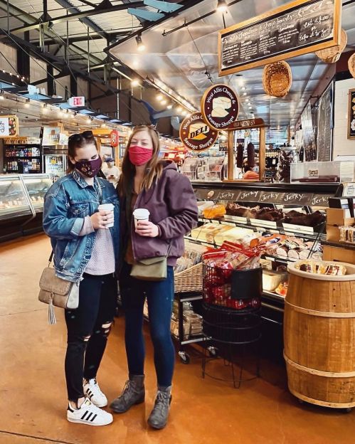 Two women stand in a market, wearing masks and holding coffee cups, surrounded by food displays and shelves.