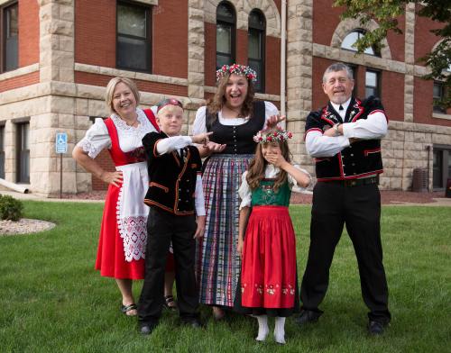 A group of five people in traditional costumes, smiling and posing outdoors in front of a historic building.