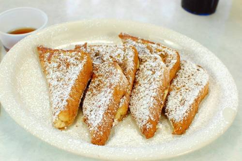 A plate of golden-brown French toast slices dusted with powdered sugar, served with a small cup of syrup.