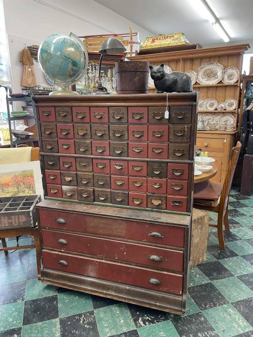 A vintage wooden cabinet with numerous small drawers, topped with a globe and a black cat figurine, in an antique shop.