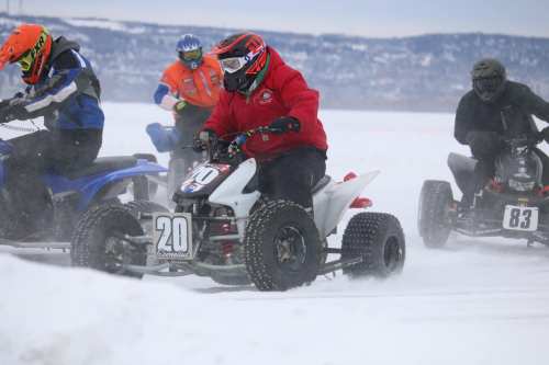 Four riders on ATVs race across a snowy landscape, kicking up snow in a winter competition.