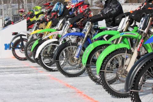 A lineup of colorful dirt bikes on ice, with riders in gear ready for a race.