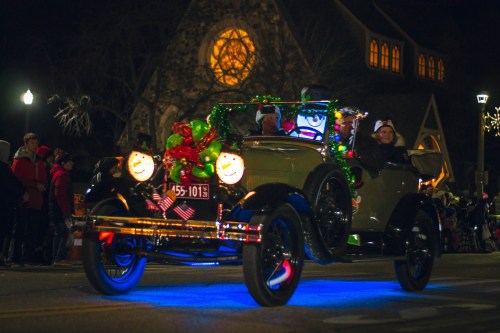 A decorated vintage car with festive lights and holiday decorations drives through a nighttime parade.