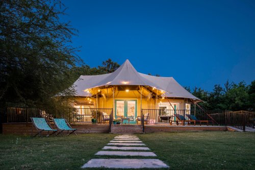 A cozy tent-style cabin with a large canopy, surrounded by greenery and a pathway, illuminated at dusk.