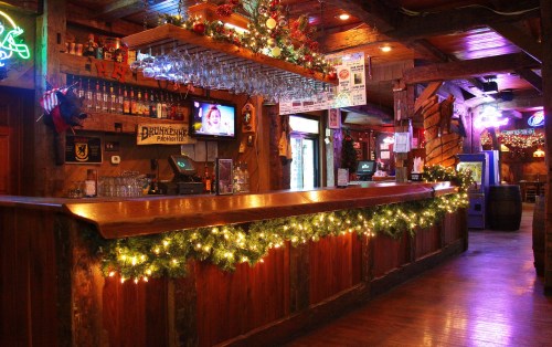 A cozy bar decorated with festive lights and greenery, featuring a wooden counter and shelves stocked with drinks.