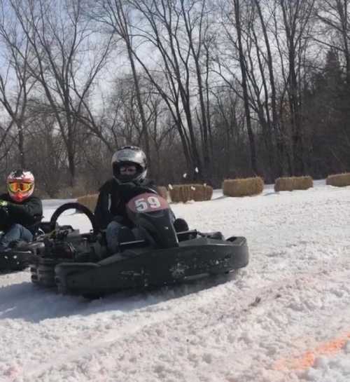 Two go-karts on a snowy track, with drivers wearing helmets and racing gear, surrounded by hay bales.