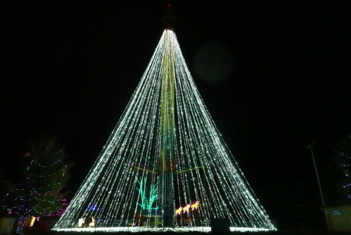 A brightly lit Christmas tree made of lights, glowing in a dark night, with colorful decorations in the background.