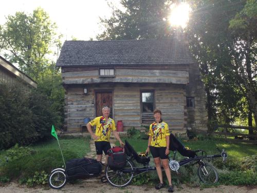 Two cyclists in matching yellow jerseys stand beside their recumbent bikes in front of a rustic log cabin.