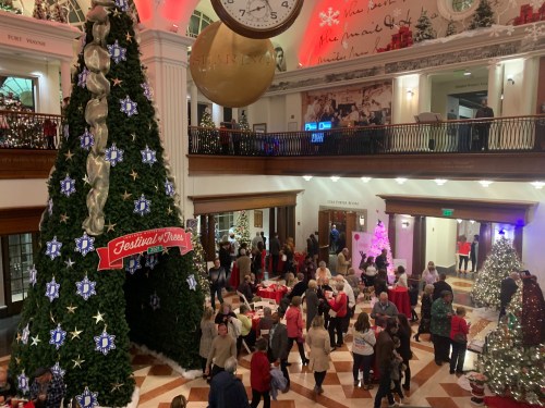 A festive indoor scene with a large Christmas tree, decorations, and a crowd enjoying a holiday event.