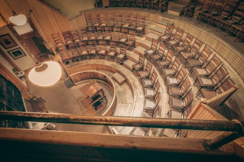 A circular auditorium with wooden chairs arranged in tiers, viewed from above, featuring a central stage area.
