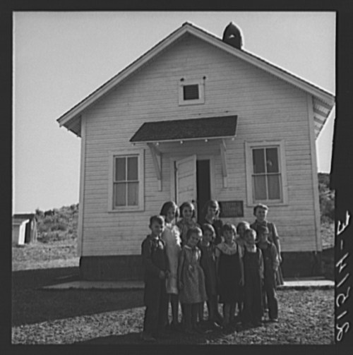 A group of children stands in front of a small, white schoolhouse on a sunny day.