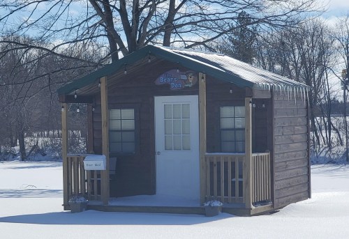 A small wooden cabin with a green roof, surrounded by snow and trees, featuring a sign that reads "Beans' Den."