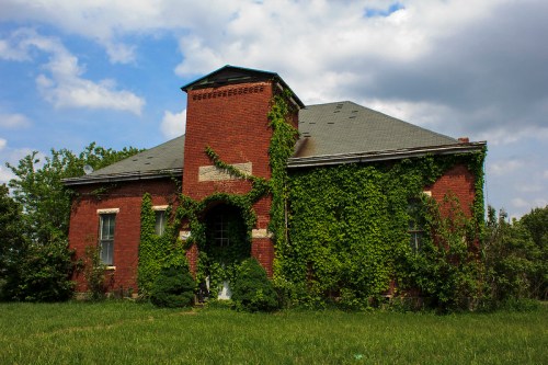 A brick building covered in green ivy, surrounded by grass and trees, under a cloudy sky.