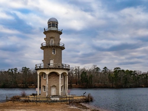 A tall, weathered lighthouse stands by a calm lake, surrounded by trees under a cloudy sky.