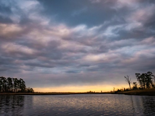 A tranquil river scene at sunset, with cloudy skies and silhouettes of trees along the water's edge.