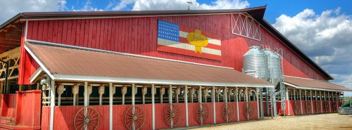 A red barn with a large mural featuring an American flag and a yellow emblem, alongside silos under a cloudy sky.