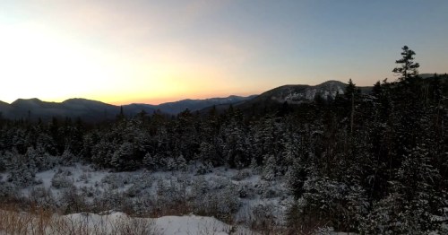 A serene winter landscape with snow-covered trees and mountains under a colorful sunset sky.