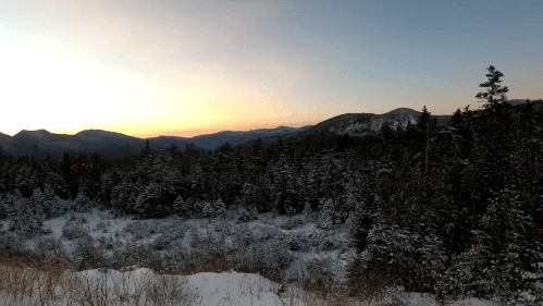 A serene winter landscape at sunset, featuring snow-covered trees and distant mountains under a colorful sky.
