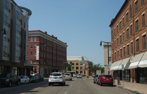 A city street lined with brick buildings and parked cars under a clear blue sky.