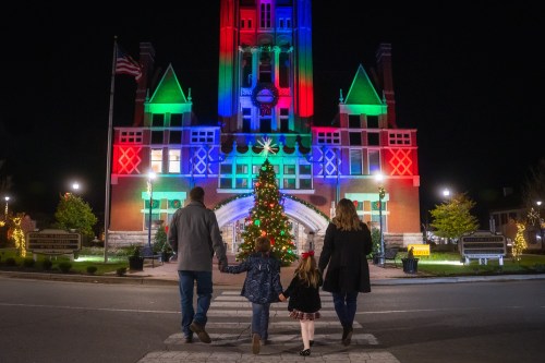 A family walks hand-in-hand past a brightly lit building and Christmas tree at night.