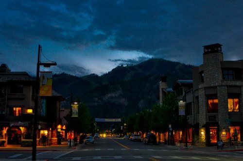 A quiet street at dusk, lined with shops and trees, with mountains in the background under a cloudy sky.